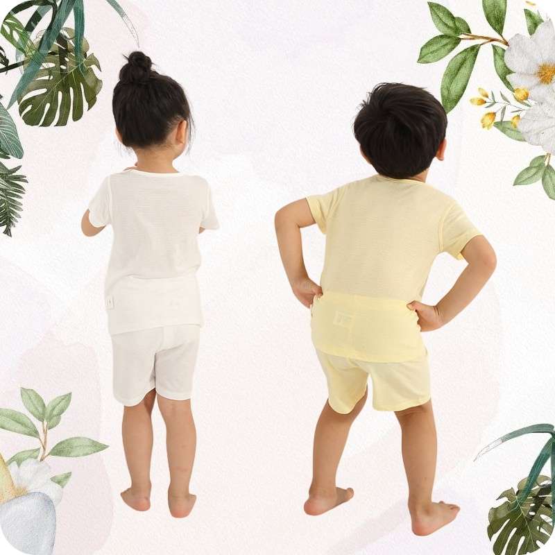 Two children wearing yellow and white outfits standing back-to-back with a floral and leafy background.