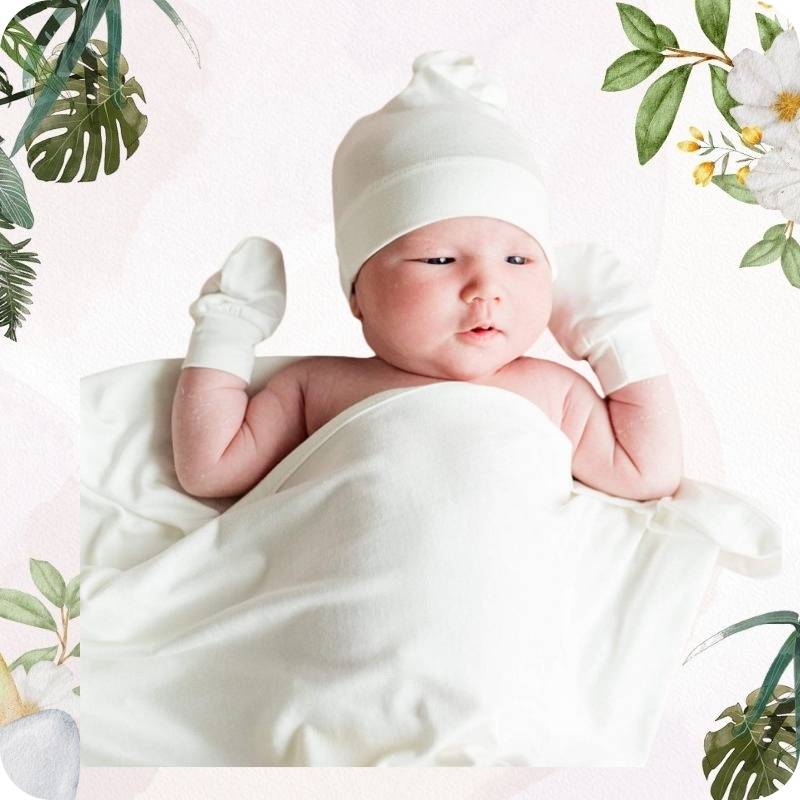 Newborn baby wrapped in white with a white hat, surrounded by green leaves and flowers.