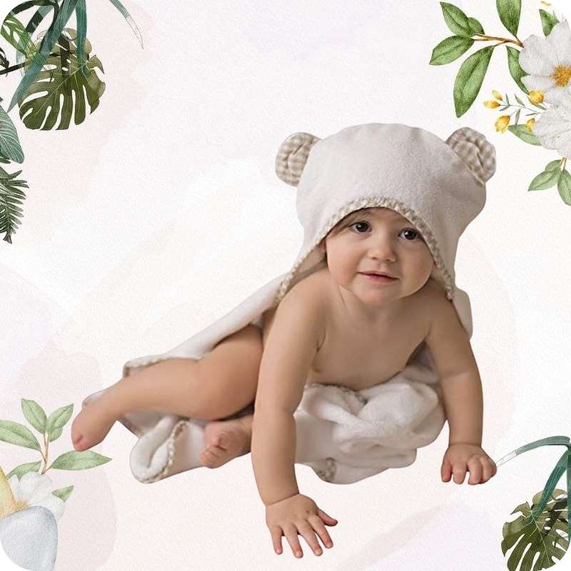 Baby wearing a white hooded towel with bear ears, surrounded by green leaves and flowers on a light pink background.